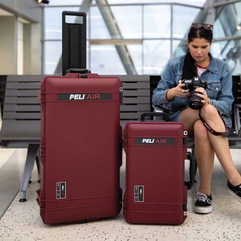 Two oxblood Peli Air Travel hard cases at an airport terminal beside a woman checking a camera while seated.