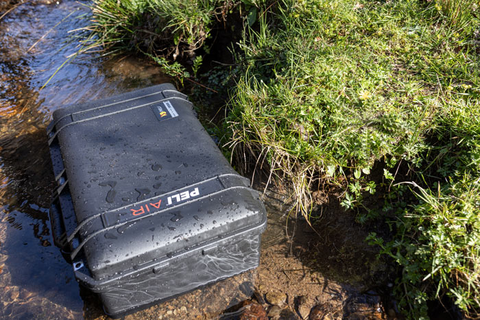 A Black 1555 Peli Air case in a shallow stream next to some grass.