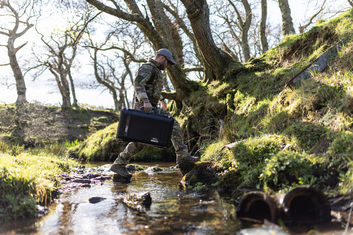 A man in camouflage clothing carrying a Peli 1555 Air case across a stream.